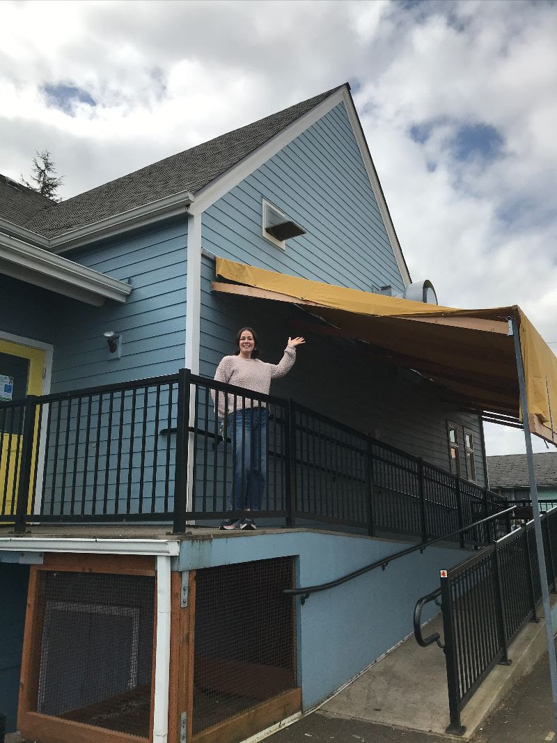 A brunette white woman waving and smiling at the camera, while standing on the porch of a large blue building